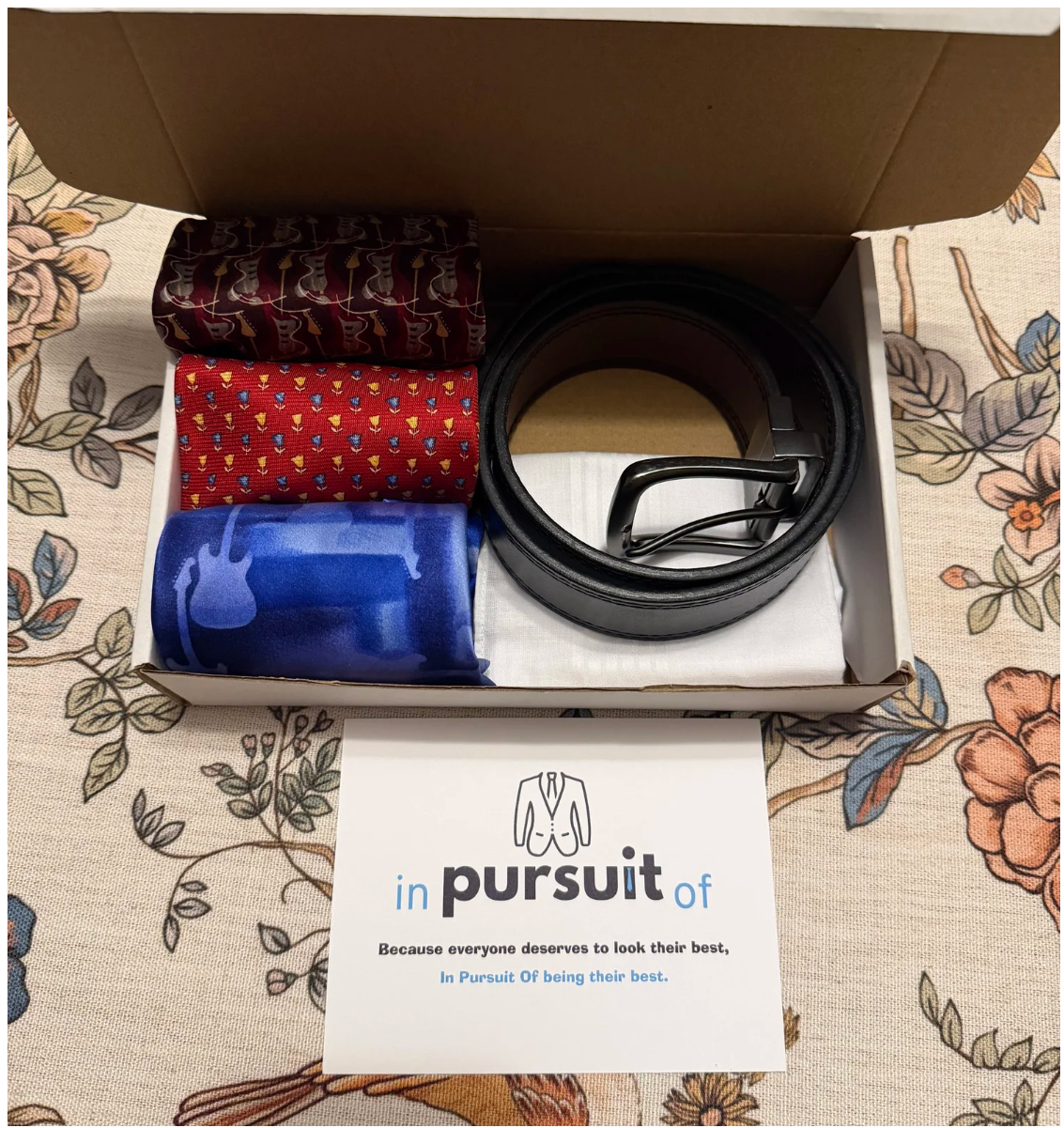 In Pursuit Of gift box containing ties, a leather belt, and a branded card reading 'Because everyone deserves to look their best, In Pursuit Of being their best.'