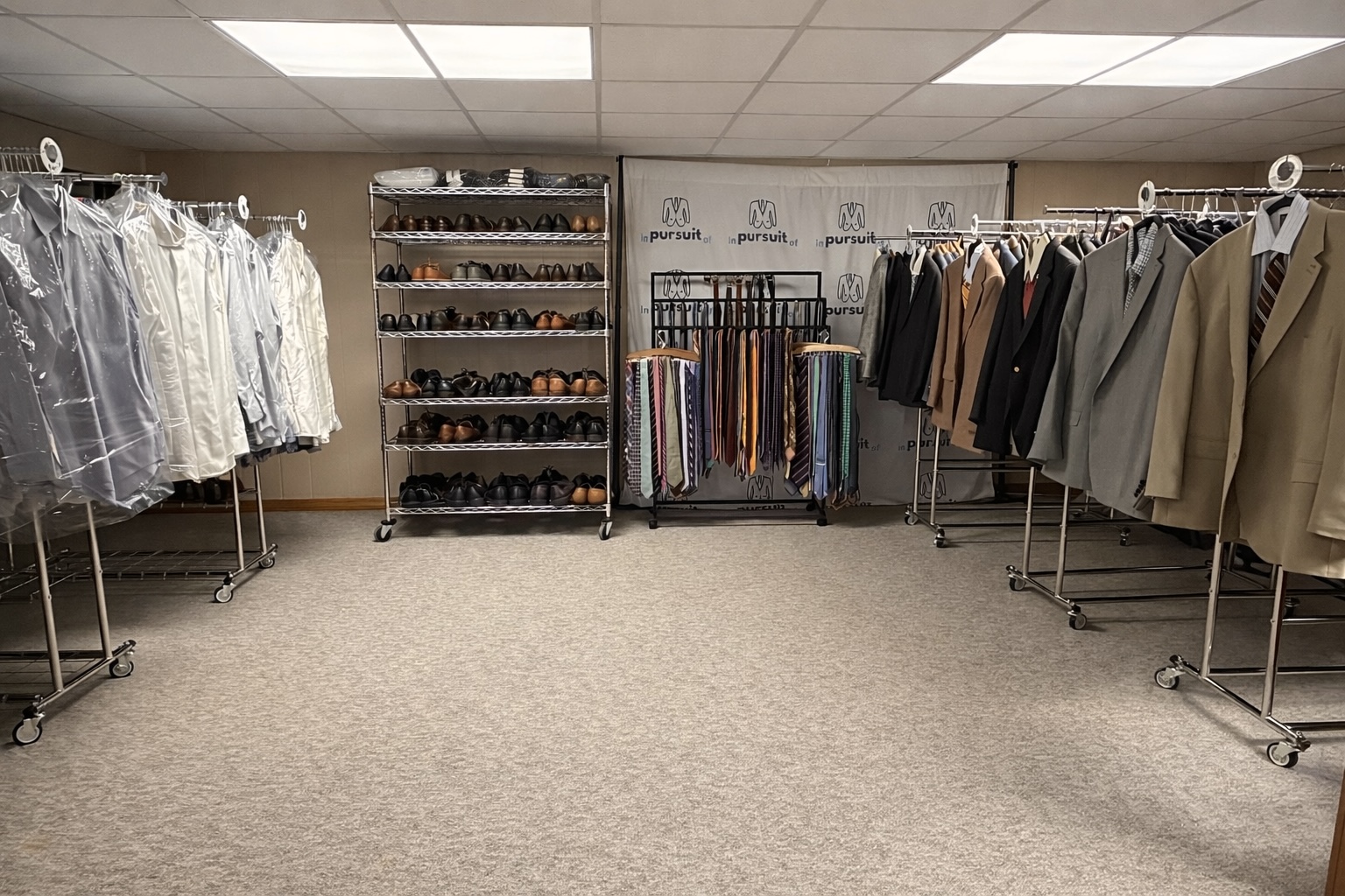 In Pursuit Of clothing inventory room with racks of donated men's business suits, dress shoes, and colorful neckties